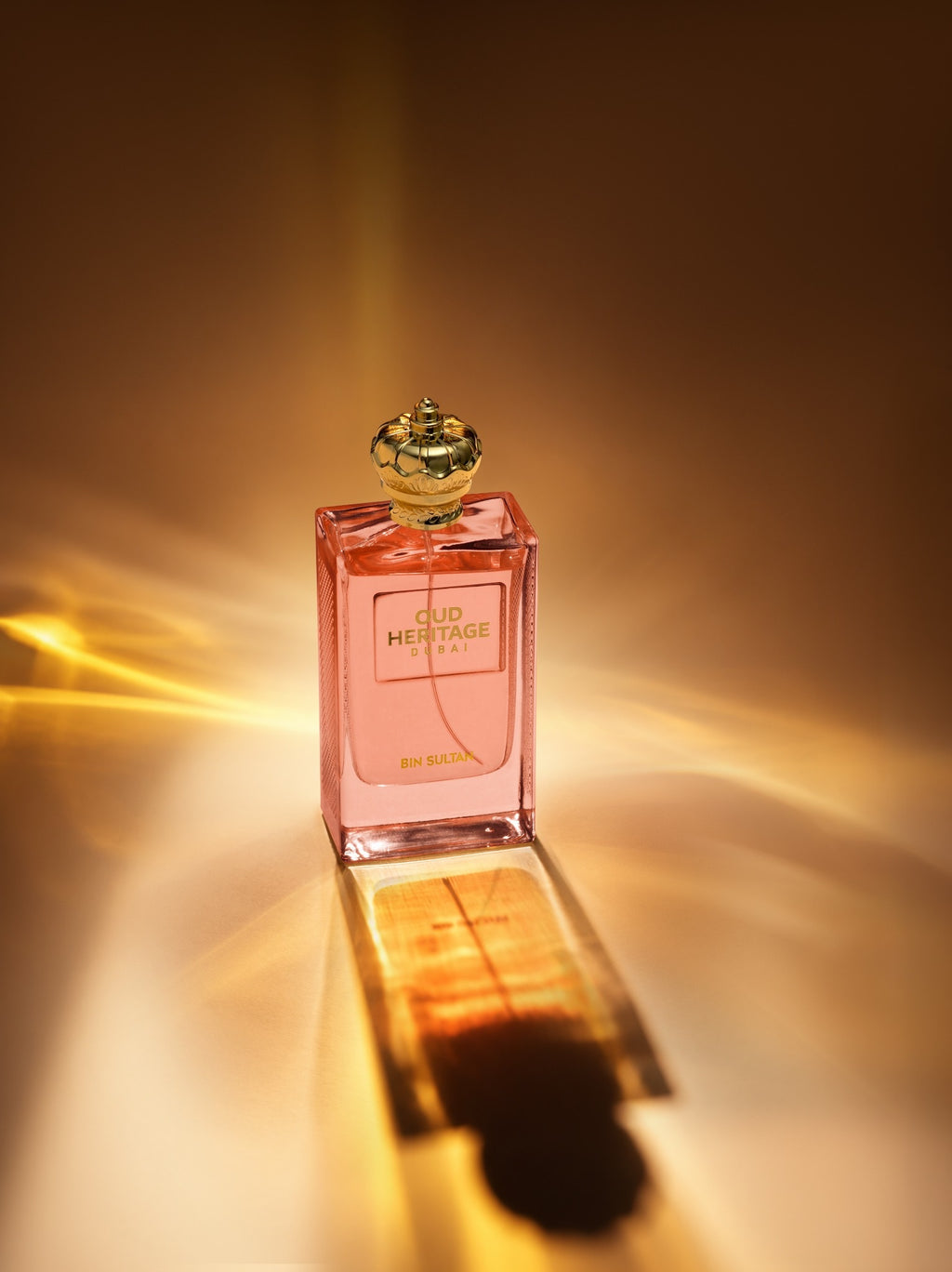 A rectangular glass perfume bottle filled with pink liquid, featuring a gold cap and a front label that reads 'OUD Heritage DUBAI' and 'En Rose'. The bottle is photographed on a reflective surface with dramatic warm light rays and a reflection against a dark brown background.