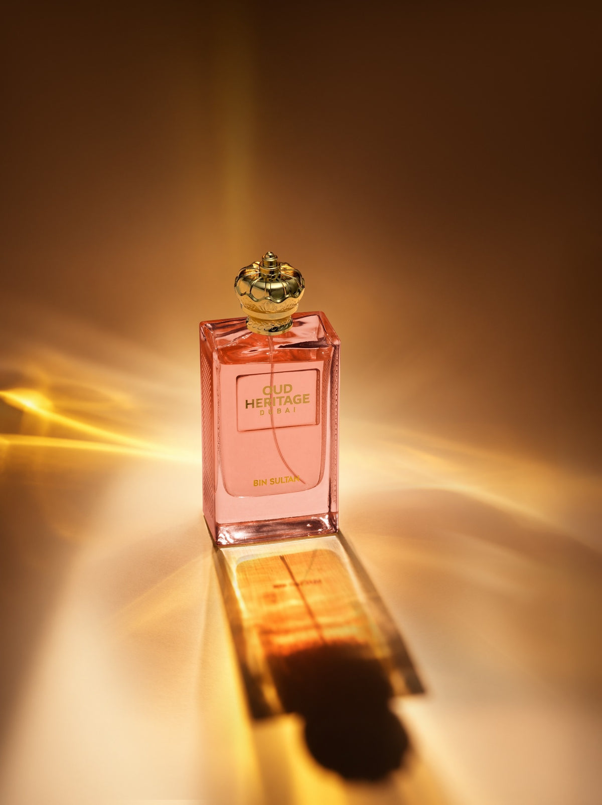 A rectangular glass perfume bottle filled with pink liquid, featuring a gold cap and a front label that reads 'OUD Heritage DUBAI' and 'En Rose'. The bottle is photographed on a reflective surface with dramatic warm light rays and a reflection against a dark brown background.