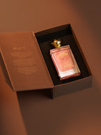 A rectangular glass perfume bottle filled with pink liquid, displayed inside an open brown presentation box with a gold cap, on a warm brown background.