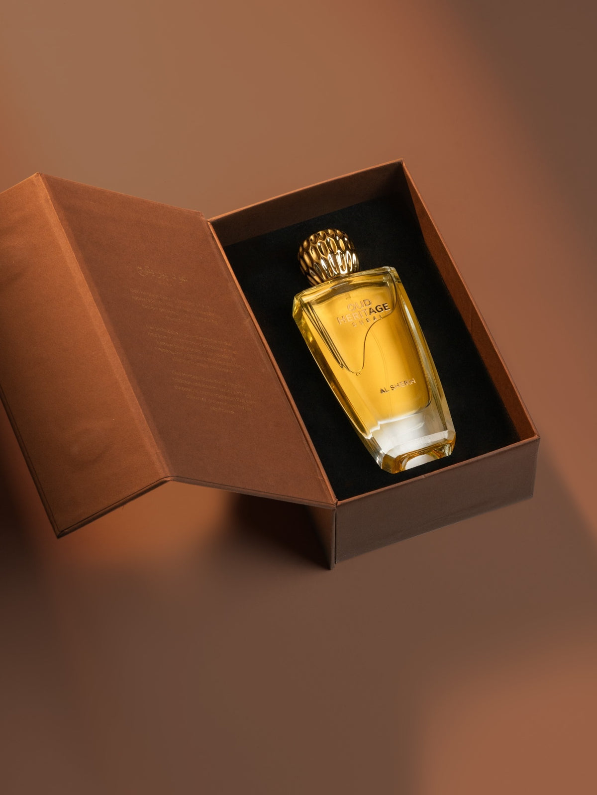 A rectangular glass perfume bottle filled with amber-yellow liquid, set inside an open brown presentation box with a black interior. The bottle has a textured gold cap and subtle gold lettering on the front.