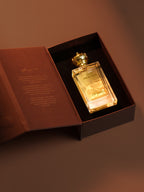 A luxury perfume bottle with a gold cap and gold lettering, presented inside an open dark brown box with a matching brown inner lid featuring gold text, set against a warm brown background.