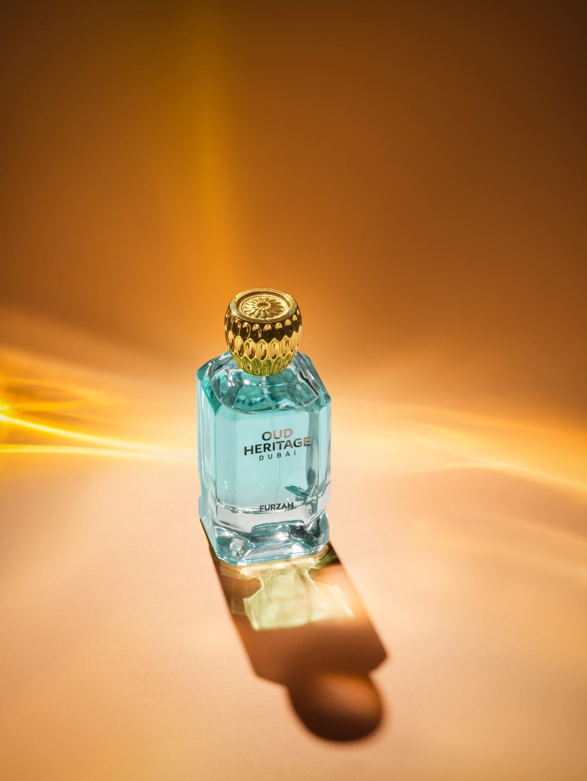 A faceted light blue glass perfume bottle with a textured gold cap, labeled 'OUD HERITAGE DUBAI', set against a warm golden-brown background with dramatic light flares and a soft reflection.